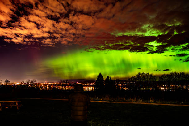 Northern Lights over a inhabited mountain with residential lights and moonlight and a man looking at the show.
