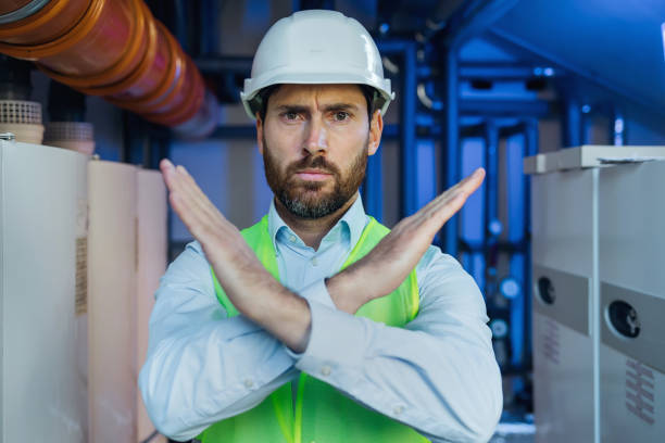istock Electrical engineer technician in protective vest and white helmet crossing arms signaling danger. Substation worker say stop, backdrop with blue light due to high voltage 2152658307