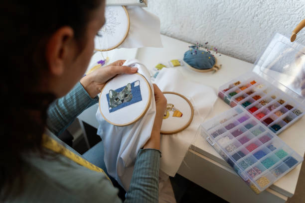 The detailed process of cross-stitch embroidery on burlap fabric. A metal embroidery needle is threaded with vibrant red thread, working on a red and white floral pattern. The red cross-stitches form a neat background while the white thread creates delicate motifs, demonstrating skillful needlework. The texture of the burlap fabric underneath adds a rustic and traditional touch to the overall composition, emphasizing the handmade essence of the craft.