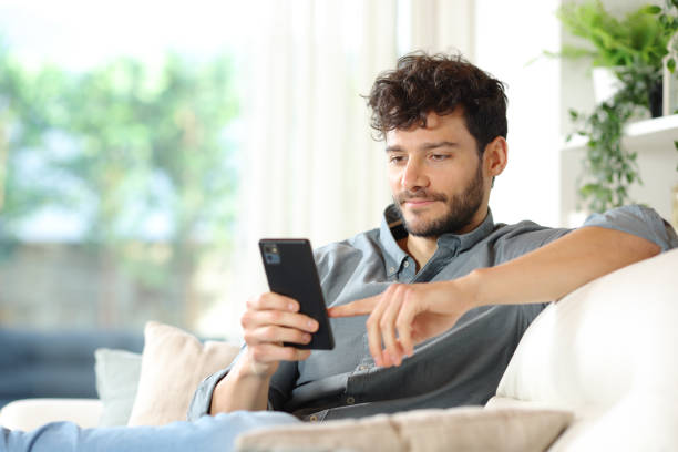 seriózní muž používající telefon na gauči doma - mladí muži - stock snímky, obrázky a fotky
