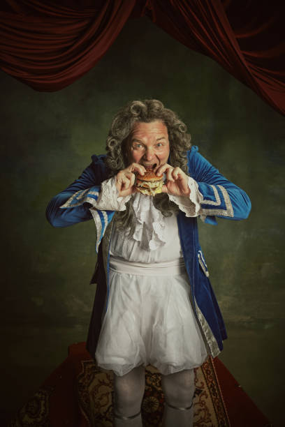 istock Hungry elderly man, dressed as king in classical baroque style clothes eating modern hamburger against vintage background. 2152704706