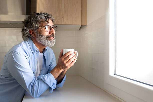 happy mature man relaxing at home having morning coffee looking out the kitchen window. copy space. - rotina imagens e fotografias de stock