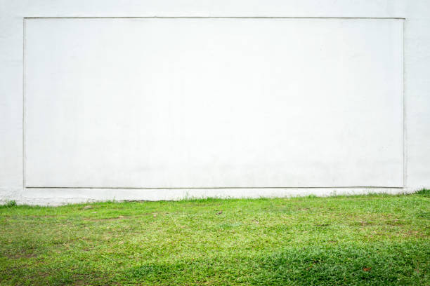Large white wall with a square frame and green grass in the foreground. Frontal view of a bright house facade as a background