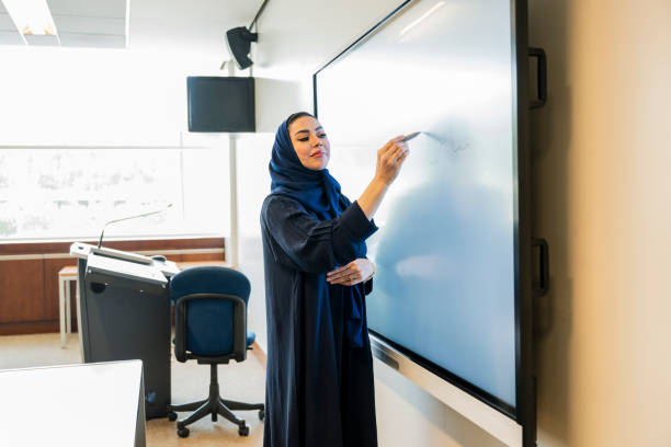 Smiling Saudi educator in traditional clothing standing at front of university classroom and augmenting her lecture with visual notes.