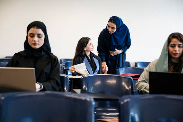 View between classmates sitting at desks and using laptops to mature educator face to face with smiling young woman getting help.