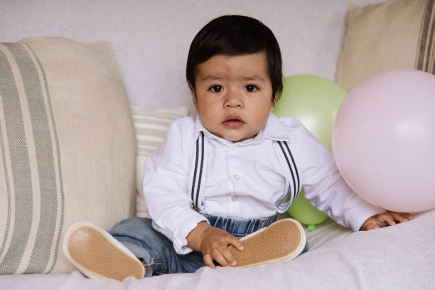 Portrait of a cute baby boy sitting on a sofa, dressed in a stylish white shirt with suspenders and denim jeans.