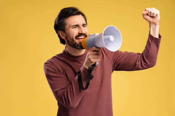 istock Happy young man shouting by megaphone, holding hand up, isolated on yellow 2152760647