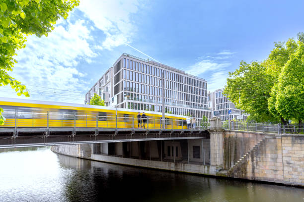 This image captures a dynamic urban scene in Berlin, featuring a yellow train speeding over a bridge, surrounded by modern architecture and lush greenery.