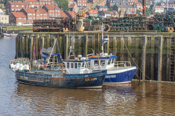Whitby, UK. May 1, 2024.  Two small fishing boats are moored alongside a quay and buildings are in the background. Lobster pots are stacked on the quayside.