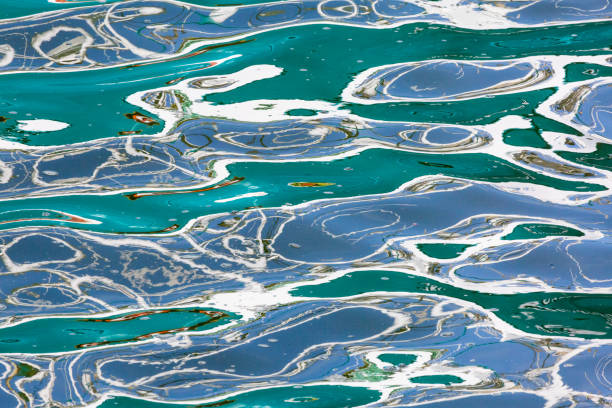 Gentle waves create intricate patterns on the surface of water varying in hues of blue and green, the colours of a fishing boat at harbour on Amble quayside on the coast of Northumberland, North East England. Reflected light dances across the ripples, intertwining with the water's natural color and creating abstract shapes.