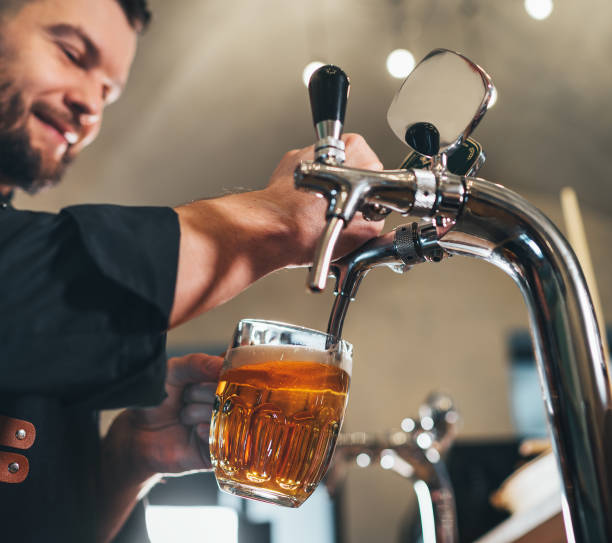istock Smiling stylish bearded barman dressed black uniform with an apron tapping fresh lager beer into glass mug at bar counter. Successful people, beer consumption, beverages industry concept image 2152820148
