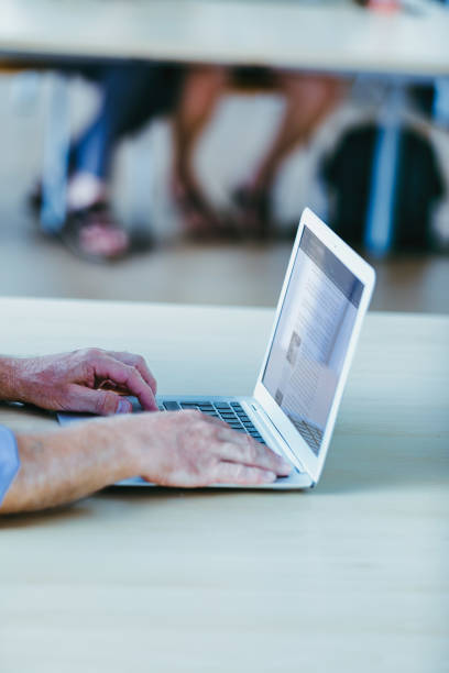 elderly man's hands type on a laptop in a bright room, with the blurred background showing legs and feet. The man wears a blue shirt, focusing on the screen in front of him