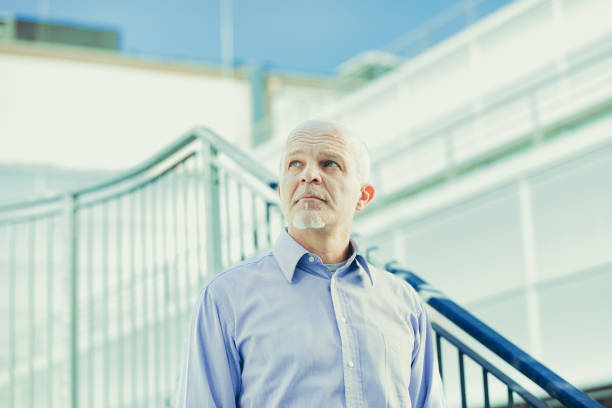 Mature man stands confidently outdoors, blue shirt enhancing his wise demeanor against a modern architectural backdrop, exuding professionalism