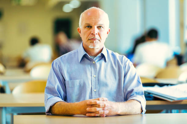 elderly man with a serious expression sits at a table in a busy, well-lit room, hands clasped. He reflects on future issues like climate change, job losses due to automation, and power concentration