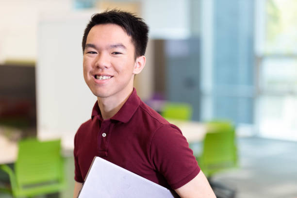 istock Happy young student standing in a classroom holding a laptop 2152833234