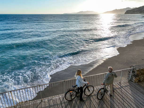Mature couple push bicycles along walkway above coastline, Liguria