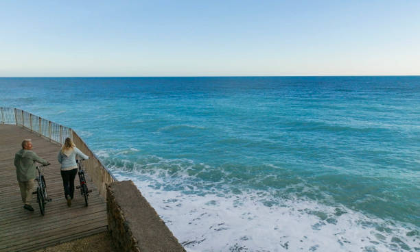 Mature couple push bicycles along walkway above coastline, Liguria