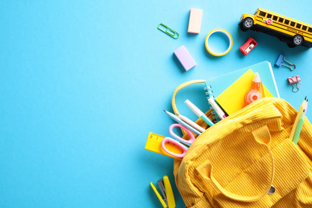 istock Yellow backpack overflowing with school essentials such as pencils, scissors, and multicolored notebooks, complemented by a miniature school bus, all set against a striking blue backdrop 2152851087