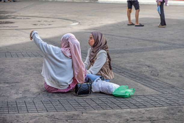 Sandakan, Eastern Borneo, Malaysia - February 12th 2024:  Two young women with hijabs sitting on the ground in a public square taking a selfie