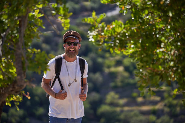 A man in a denim jacket and hoodie hikes through an autumn forest, carrying binoculars and a backpack, smiling as he strides along with trekking poles in a scenic outdoor setting.
