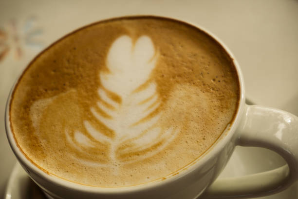 An elegantly presented latte sits in a classic ceramic coffee cup, featuring intricate latte art on its creamy milk foam surface. The warm, inviting tones of the coffee blend beautifully with the milky white, creating a visually appealing contrast. Set against a softly blurred background, this image captures the artistry and appeal of a freshly brewed cup of coffee. Perfect for use in cafes, beverage menus, or any project emphasizing the art of coffee making.