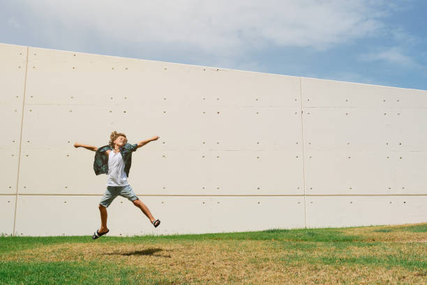 Full body of energetic boy in casual clothes jumping above ground with raised arms on green lawn against wall