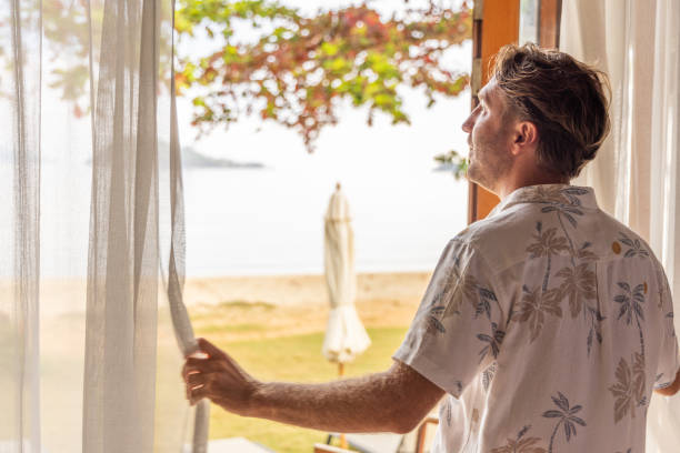istock Man Opening Sheer Curtains to a Beach Scene 2152868964