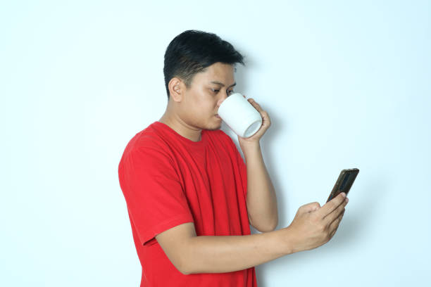 istock Side view of a young Asian man drinking while looking at a smartphone. Wearing a red t-shirt 2152870168