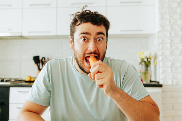 Front view looking at camera young man sits at a table and eats raw carrot with his eyes wide open.