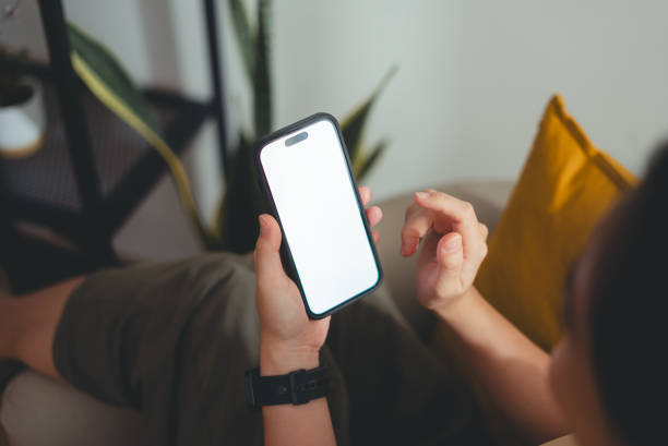 Woman holding smart phone with white screen mock-up sitting on armchair in living room at home.