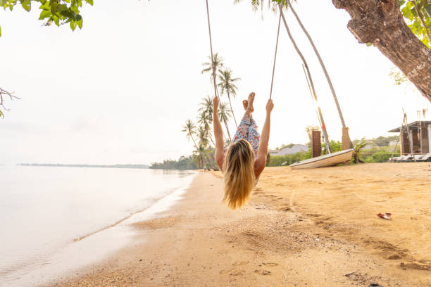 A woman swings joyfully on a tree swing by a sandy beach with her legs in the air, she's surrounded with palm trees and an ocean in the background, enjoying a beautiful tropical sunset.