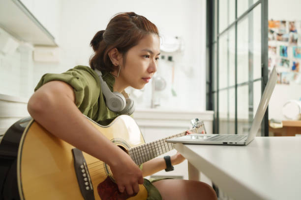 istock Focused Asian Woman Learning Guitar Through Online Tutorial on laptop computer, Engaged in Musical Practice in Kitchen 2152898646