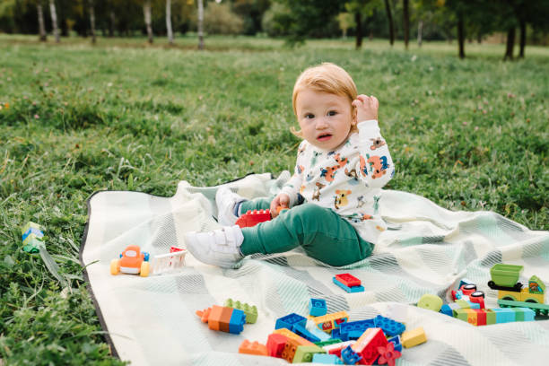 Toddler boy little cute walking and playing small car toys in garden. Kid 2 year old play constructor game sitting in green grass on blanket in park sunny summer day. Outdoors creative activities. niño pequeño lindo caminando y jugando juguetes pequeños de coche en el jardín. niño de 2 años juega al juego de constructor sentado en la hierba verde sobre una manta en el parque día soleado de verano. actividades creativas al aire libre.