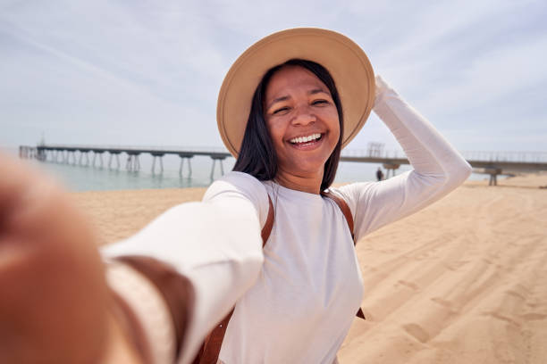 istock African woman using cellphone to take a selfie standing on the sand beach, looking at camera smiling 2152904900