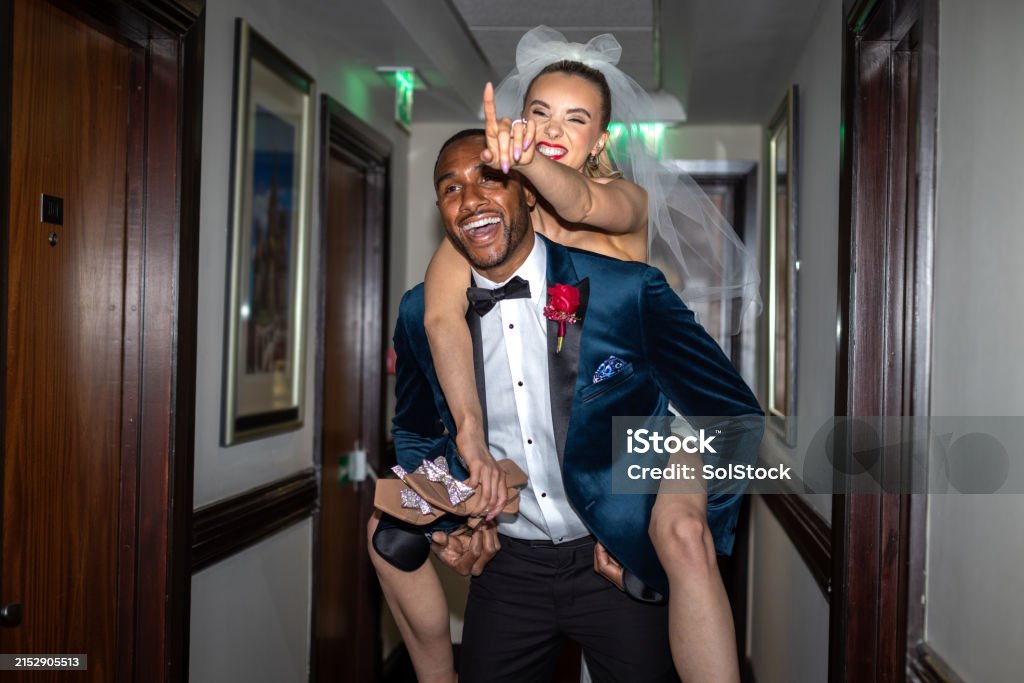 Pointing The Way Medium shot of a newlywed couple in a hotel corridor. The young bride is piggybacking on her husband's back. She is laughing while pointing in the direction of the hotel room and carrying her high heels. The groom is carrying her while smiling.Videos are available similar to this scenario. Wedding Stock Photo Pointing The Way Medium shot of a newlywed couple in a hotel corridor. The young bride is piggybacking on her husband's back. She is laughing while pointing in the direction of the hotel room and carrying her high heels. The groom is carrying her while smiling.Videos are available similar to this scenario. Wedding Stock Photo
