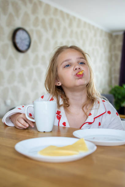 A charming blonde baby in cute pajamas is sitting at a comfortable table in the kitchen and enjoying a delicious and healthy breakfast. The idea of a proper and balanced diet.