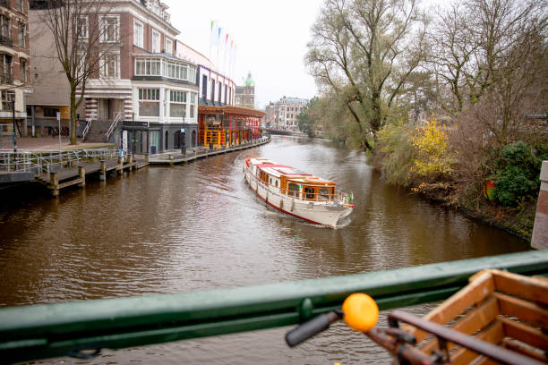 Amsterdam, Netherlands - August 8, 2024: Amsterdam North Holland The Netherlands Europe, Dutch Building Exterior, Parking Lot, Land Vehicle, Tourists Sitting Down Inside Amsterdam Canal Sightseeing Recreational Boat, Locals Walking Around, Eating And Drinking Scenery During Summer Season