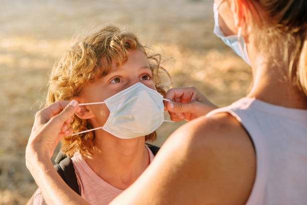 Unrecognizable woman putting protective mask on preteen boy with rucksack while standing in nature during COVID 19 pandemic