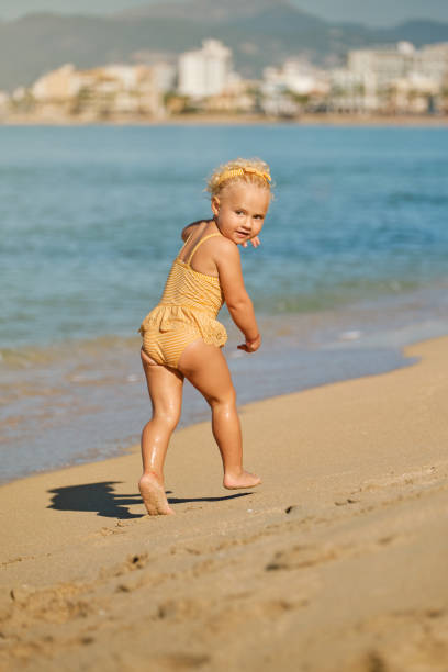 Back view of carefree toddler in yellow swimsuit and headband having fun while running along sandy seashore and looking over shoulder on summer day