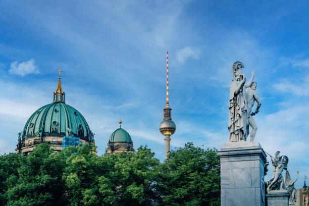 Capturing the juxtaposition of Berlin Cathedral's historic dome with the modern TV Tower. Statues in foreground under a clear blue sky. Architectural contrast and urban exploration in Germany.