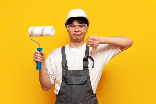 istock dissatisfied asian guy painter in uniform holds roller and shows dislike on yellow isolated background 2152958219