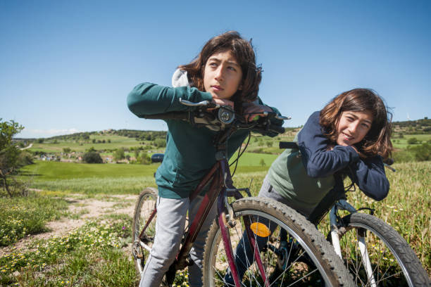 Two young brothers ride their bicycles in a field. Both are resting on the handlebars of their bicycles.