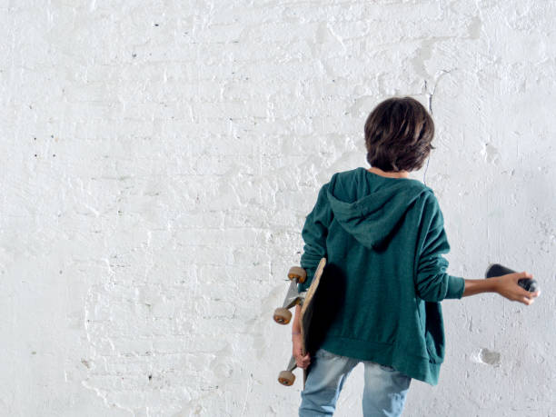 A boy is holding a skateboard and a spray paint for graffiti. He is wearing a green hoodie. Concept of youthfulness and creativity, as the boy is likely to be interested in skateboarding and street art.