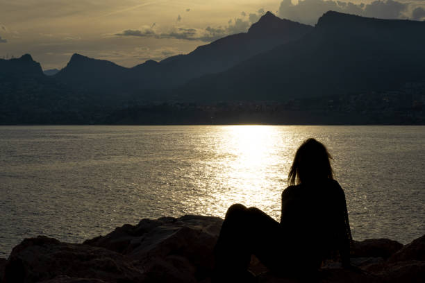This striking image captures the silhouette of a woman in a moment of peaceful contemplation. Seated on the rugged terrain, she gazes out at the tranquil waters, with the sunset behind distant mountains casting a golden reflection upon the scene