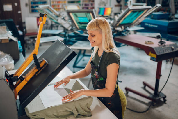 Smiling printing workshop worker using heating press and transfer on tee Smiling female printing workshop worker using transfer on a heating press at facility. Happy female graphic technician standing at printing shop and using heating press for transferring. Copy space. t shirt transfer stock pictures, royalty-free photos & images