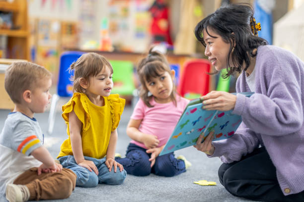 Reading is Fun! A preschool teacher sits on the floor of her classroom with a small group of students as she reads them a book. The children are each dressed casually and are focused on the story. child care stock pictures, royalty-free photos & images