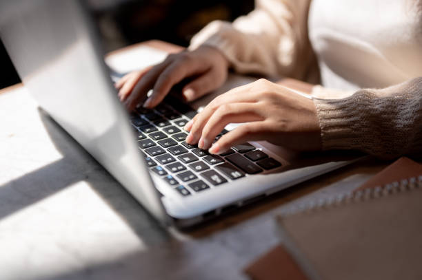 close-up image of a woman typing on keyboard, working on her laptop computer at a table indoors. - dactilografar imagens e fotografias de stock