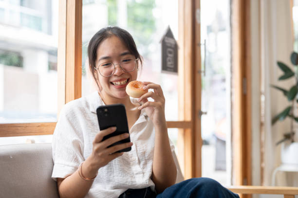 istock A young, happy Asian woman enjoying a donut while using her smartphone on a sofa in a cafe. 2153012339