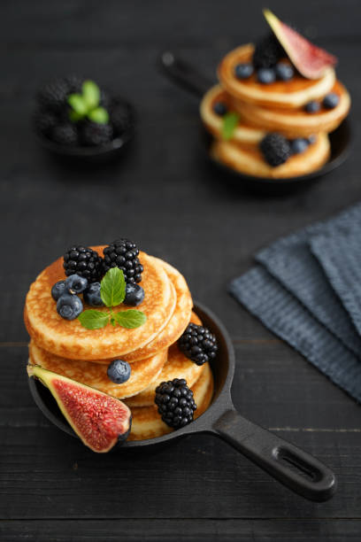 Image focuses on first pan with golden pancakes,  contrasting against a black wooden background. Adorned with vibrant blueberries, fig, blackberry and green vibrant mint leaves.
