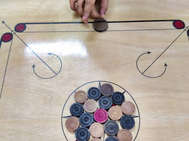 Picture of a carrom board with carrom coins arranged in center of the board and a person with a striker kept at one end of the board